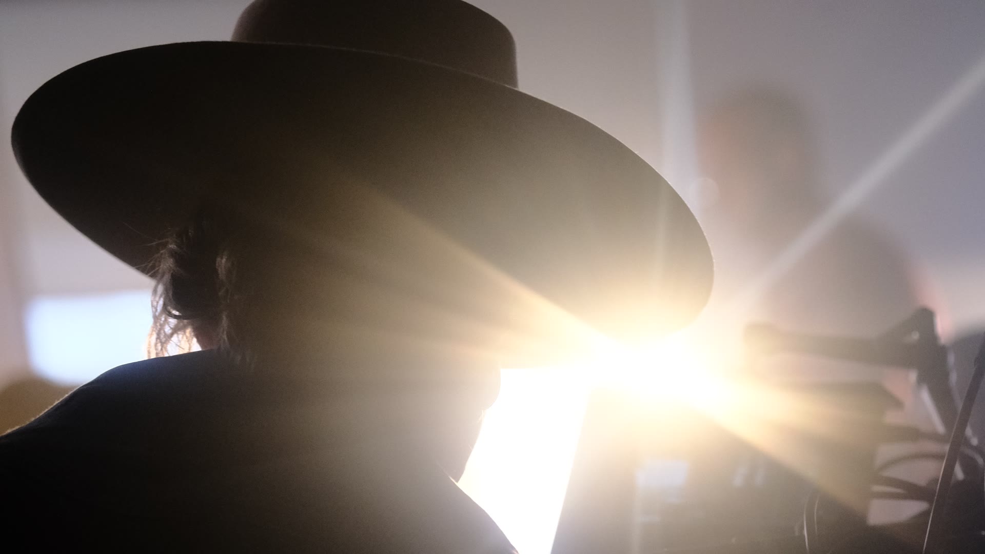 Angelia Hughes silhouetted in her flat-brim hat beside a camera, backlit by a lamp flaring into the lens.